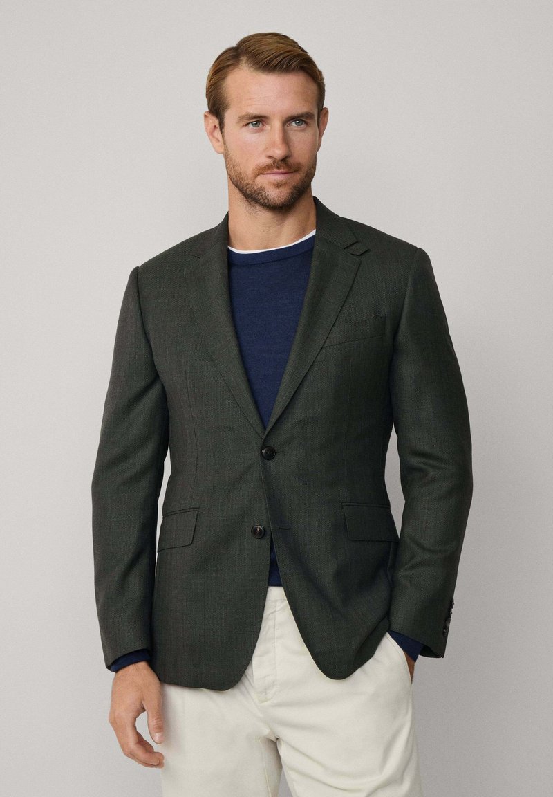 Tmavozelený sako s textúrovanou tkaninou, s jednoradovým zapínaním, dvoma gombíkmi a klopami, spárované s námorníckym svetrom a krémovými nohavicami.