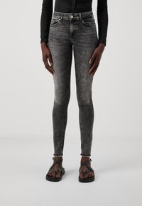Jeans de mezclilla negra ajustados con un lavado desgastado, cinco bolsillos y costuras en contraste. Combinados con unas sandalias negras de tiras con suela plana.