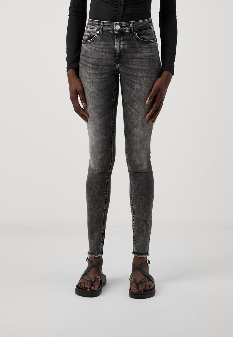 Jeans de mezclilla negra ajustados con un lavado desgastado, cinco bolsillos y costuras en contraste. Combinados con unas sandalias negras de tiras con suela plana.