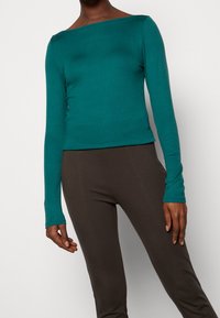 Teal långärmad topp med vid halsringning, figurnära design och mjuk textur, kombinerad med mörkbruna figurn ära byxor med synlig sömnad.