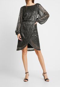 Schwarzes Kleid mit metallisch plissierter Textur, langen transparenten Ärmeln, einem Bootsausschnitt und einem knielangen Saum mit diagonaler Schnittführung. Kombiniert mit schwarzen Absätzen.