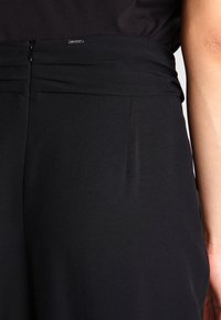 Svarta högmidjade byxor med plisserad design. Slät tygkvalitet, diskret dragkedjeöppning bak och sidofickor.