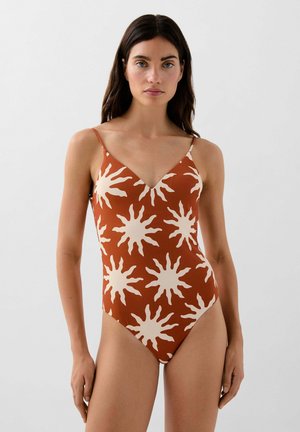 Mujer con cabello largo y oscuro lleva un traje de baño enterizo color óxido con grandes patrones blancos de rayos de sol, de pie contra un fondo claro liso.