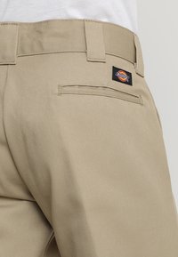 Detailní záběr béžových kalhot Dickies, ukazující zadní kapsu, poutka na opasek a logo na pravé straně.
