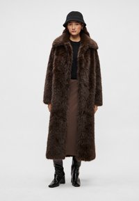 Hnedý kabát z umelého kožušiny so širokým golierom, dlhými rukávmi a priamym strihom, skombinovaný s čiernym topom a hnedou sukňou.