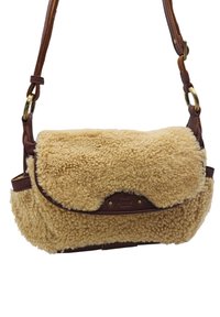 Sac en shearling beige avec des accents en cuir marron, un rabat repliable, une bandoulière ajustable et des détails en laiton. Design compact et texturé.