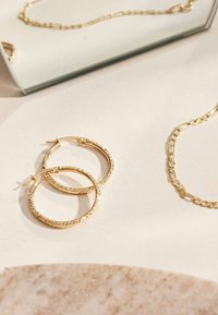 Gouden hoepel oorbellen met een gestructureerd, patroon oppervlakte, vergezeld van een delicate gouden kettingketting op een lichte ondergrond.