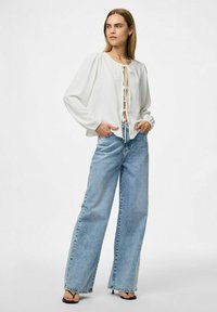 Weiße Bluse mit langen Ärmeln, rundem Ausschnitt und Bindeband vorne, kombiniert mit hellblauen weit geschnittenen Jeans.