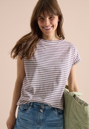 Jeune femme souriante, portant un t-shirt rayé violet et blanc, un jean bleu, tenant un sac matelassé vert clair avec une doublure à motifs.