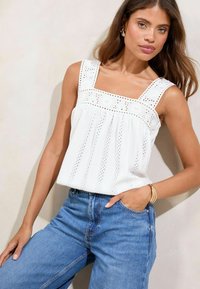 Haut sans manches blanc avec décolleté carré, détails brodés et design plissé. Associé à un jean taille haute bleu clair.