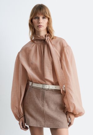 Peach-colored, lightweight blouse with a high neck tie and voluminous sleeves, paired with a beige belt and a textured brown mini skirt.