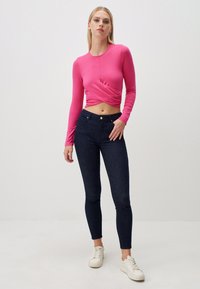 Maglietta rosa aderente a maniche lunghe con design frontale arricciato, abbinata a jeans skinny blu scuro e sneaker bianche. Look semplice e contemporaneo.