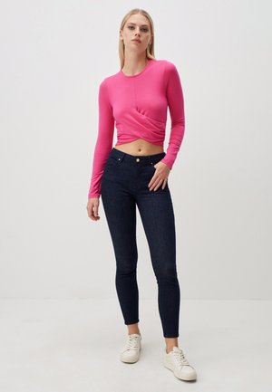 Maglietta rosa aderente a maniche lunghe con design frontale arricciato, abbinata a jeans skinny blu scuro e sneaker bianche. Look semplice e contemporaneo.