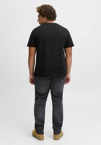 Schwarzes T-Shirt mit kurzen Ärmeln und Rundhalsausschnitt, kombiniert mit dunklen Jeans und hellbraunen Arbeitsschuhen. Die Rückansicht zeigt den lockeren Schnitt und das Design.
