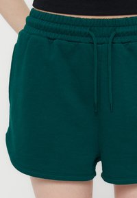 Κοντινό πλάνο ατόμου που φοράει σκούρα πράσινα ελαστικά σορτς με κορδόνι και τσέπες, σε συνδυασμό με μαύρο κοντό τοπ.