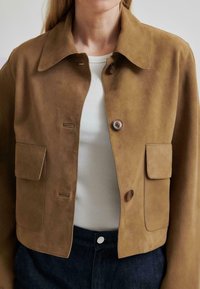 Barna velúr dzseki, inggallérral, két elülső zsebbel és egy gombos zárással. Fehér bordázott felső és sötét farmernadrág fölött viselve.