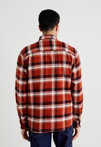 Camisa de franela a cuadros rojos y negros con mangas largas, puños abotonados, un cuello clásico y un dobladillo quecurva ligeramente en la parte de atrás.