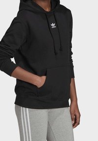Svart hoodie i bomull med en framficka, justerbar dragsko på huvan, och vit Adidas-logotyp på bröstet. Kombinerat med grå byxor.
