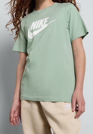 Osoba w jasnozielonej koszulce Nike i beżowych spodniach z logo Nike, stojąca na jednolitym jasnoszarym tle.