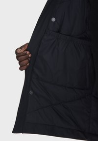 Intérieur de la veste noire montrant une doublure matelassée avec une taille élastiquée. Comprend des boutons-pression et une texture lisse et mate.