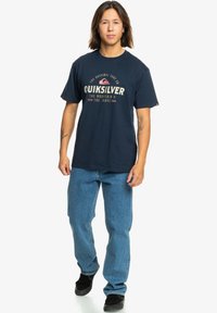 Navy blue cotton T-shirt with a round neck and short sleeves, featuring a cream and pink Quiksilver graphic logo and text. Light blue jeans.