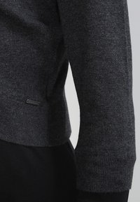 Mörkgrå stickad tröja med ribbad mudd och fåll, mjuk textur, med en liten etikett på sidan nära botten.