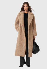 Beige oversized Mantel mit einer strukturierten, flauschigen Optik, breiten Revers, langen Ärmeln und geradem Schnitt, kombiniert mit schwarzen Jeans und High Heels.