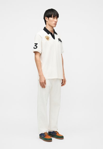 Νεαρός άνδρας που φοράει λευκό πόλο με μαύρο γιακά, λευκό παντελόνι και πολύχρωμα καφέ, πράσινα και μπλε παπούτσια, στέκεται μπροστά σε απλό φόντο.