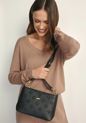 Uśmiechnięta kobieta o brązowych włosach ma na sobie beżowy sweter i trzyma czarną, wzorzystą torebkę na pasku z metalowymi, złotymi elementami.