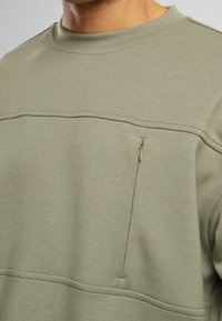 Sweatshirt vert olive en tissu doux, doté d'une petite poche zippée sur la poitrine et d'un design épuré et simple avec des détails de coutures.