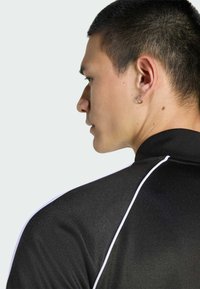 Νεαρός άνδρας με κοντά μαλλιά που φοράει μαύρο πουκάμισο με λευκές ρίγες, απεικονίζεται σε προφίλ στ' αριστερά, φορώντας μικρό σκουλαρίκι κρίκου στο αριστερό αυτί.