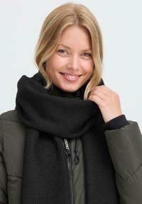 Mustrailine must sall, mis on mähitud tumerohelise polsterdatud jope ümber, millel on tõmbluku detail, millel on bränditud tõmblukktapp.