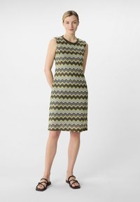 Sukienka bez rękawów w oliwkowej zieleni z wzorem w zygzaki w odcieniach beżu i brązu. Miękka tekstura, do kolan, połączona z czarnymi sandałami na cienkich paskach.