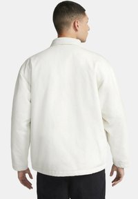 Chaqueta blanca acolchada con cuello puntiagudo, mangas largas y textura suave, con un corte relajado y un dobladillo recto en la parte posterior.