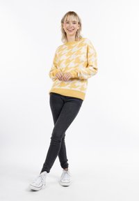Sárga és fehér houndstooth mintás kötött pulóver, bordás nyakkal és mandzsettákkal, fekete skinny farmerral és fehér sportcipővel párosítva.