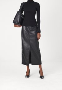 Kvinna i svart polo, svart läder midikjol med sprund framtill, svarta högklackade skor och bär en svart läder tote-väska över ena axeln.