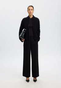 Crna pletena jakna s patentnim zatvaračem, teksturirana tkanina; crni top i široke hlače; drži crnu strukturiranu torbicu; stilizirano s visokim potpeticama.