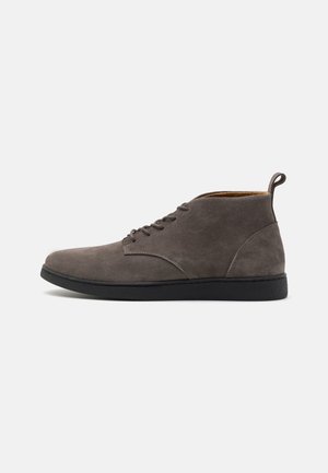 Bottine en daim gris avec semelle noire, bout arrondi, lacets à l’avant et languette à l’arrière, vue de profil sur fond blanc.