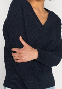 Granatowy sweter wykonany na drutach z dekoltem w kształcie litery V, ozdobiony teksturowanym wzorem w diamenty oraz ściągaczami przy rękawach i u dołu.