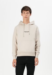 Beige Kapuzensweatshirt mit Kängurutasche und gesticktem "Originals Studio"-Logo auf der Brust, aus einem weichen Stoff gefertigt.
