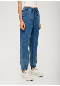 Modré denimové jogger kalhoty s elastickým pasem, bočními kapsami a volným střihem. Manžety jsou nařasené pro zúžený vzhled. Nosené s bílými teniskami.