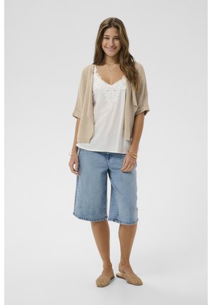 Tânără femeie în picioare, purtând un cardigan bej, un top alb cu dantelă la margine, pantaloni scurți de denim albastru deschis până la genunchi și balerini bej împletiți.