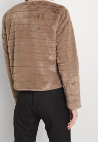 Bruine faux fur jas met horizontale strepen, ronde halslijn en geribbelde manchetten. Gedragen over een zwarte broek, met een zachte textuur.