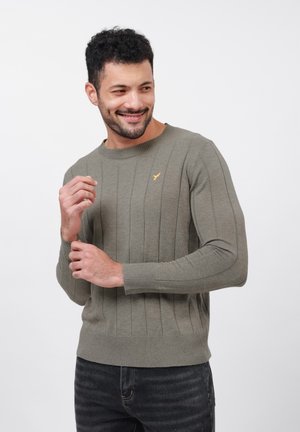 Mann, der lächelt, während er den Ärmel eines olivgrünen gerippten Pullovers mit kleinem gelben Vogel-Logo anpasst, trägt eine dunkle Jeans, vor einem weißen Hintergrund.
