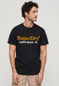 Czarny bawełniany t-shirt z krótkimi rękawami, z dużym żółtym i białym nadrukiem logo, na którym widnieje napis "SuperDry Venice Beach. CA."