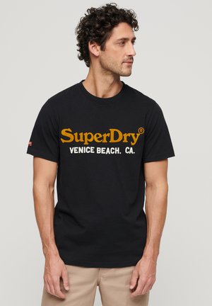 T-shirt en coton noir à manches courtes, arborant un grand logo imprimé jaune et blanc avec la mention "SuperDry Venice Beach. CA."