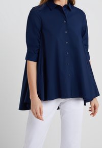 Mörkblå knapp-up skjorta med lös passform, med rullade ärmar och rundad nederkant. Tillverkad av mjukt tyg, ihopparad med vita byxor.