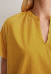 Gelbes, leichtes Shirt mit V-Ausschnitt und kurzen Ärmeln. Glatte Textur mit dezenten Nähten. Minimalistisches Design.