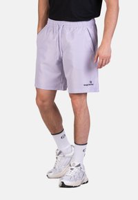 Shorts sportivi viola chiaro con vita elastica, tasche laterali e logo Sergio Tacchini sul lato sinistro in basso. Indossati con calze bianche e sneaker.