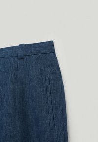 Gros plan sur un pantalon en denim bleu avec une ceinture, un passant de ceinture et une poche latérale verticale sur un fond uni clair.